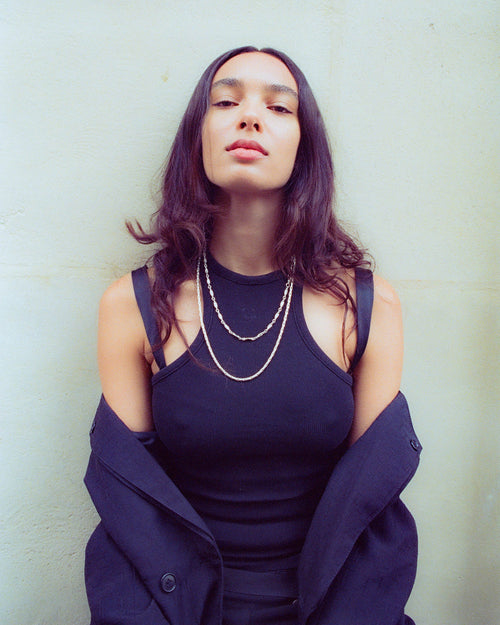 Anissa leaning on a white concrete wall wearing MAPLE 14K Gold neckalces