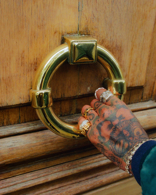 Tattooed hand wearing MAPLE jewelry grabbing large gold circular door handle on old Parisian door