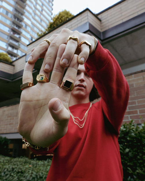 Zane Coppard in red sweater in front of brick building with the right hand palm forward and the left clasped atop pulling back fingers in 14K Gold jewelery